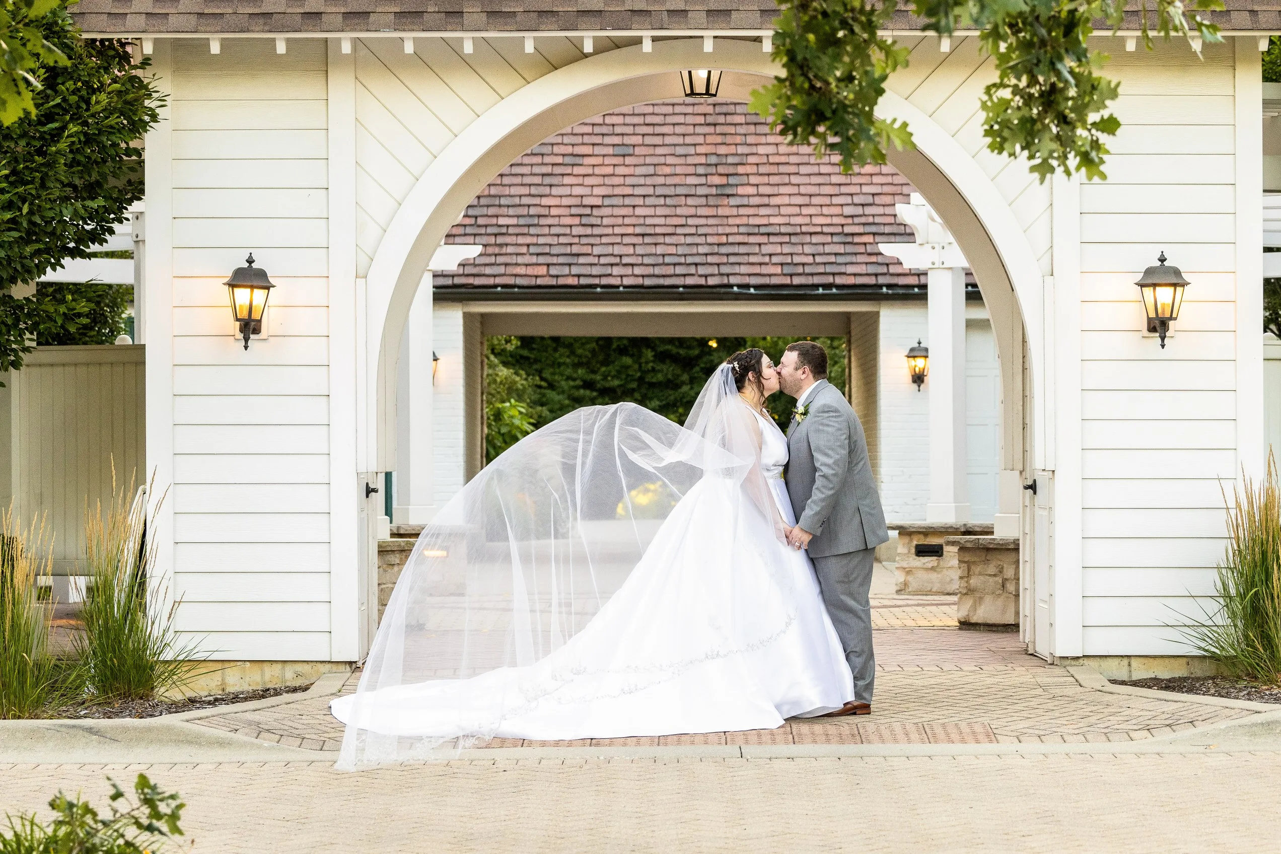 Bride and groom posing under a white wedding arch in the gardens of Danada House.
